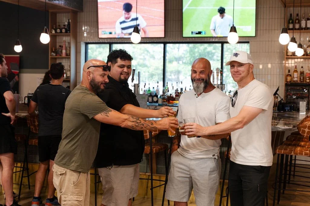 Four men raising glasses of beer in a toast at a bar, with TVs showing sports in the background.