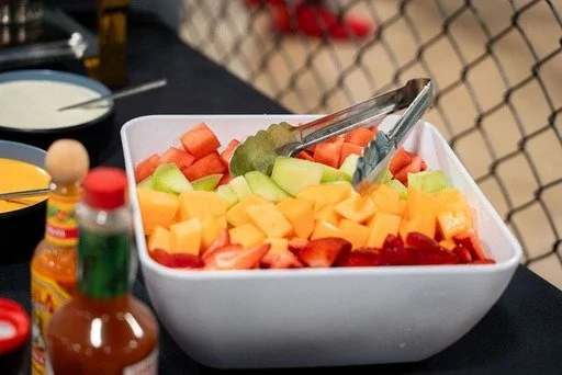 A corporate event set up of fresh mixed fruit with slices of watermelon, cantaloupe, honeydew, and strawberries, with tongs resting on the bowl, set on a black table.