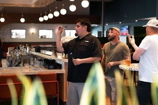 Three men at a bar or restaurant counter, one of whom is a bartender with a black shirt, and the other two are patrons, one holding a glass of beer, engaging in conversation or discussion.