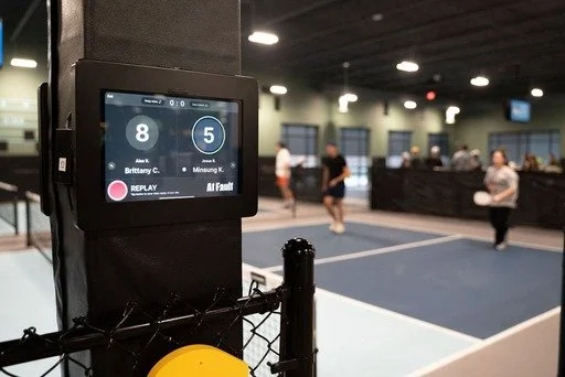 Indoor pickleball court with a digital scoreboard showing score 8-5, surrounded by players and spectators in the background.
