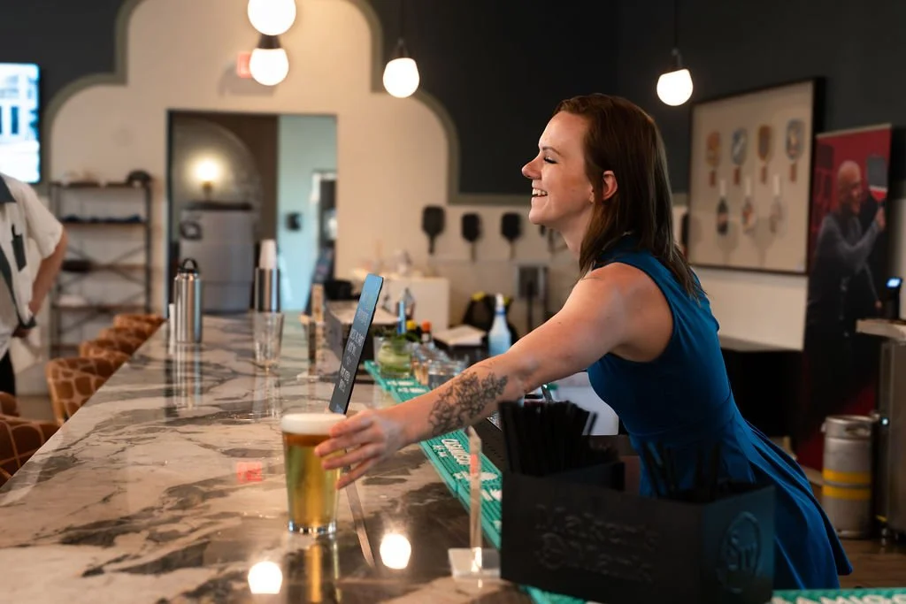 A smiling woman in a blue dress serving a glass of beer at a bar with a marble countertop and hanging lights in the background.