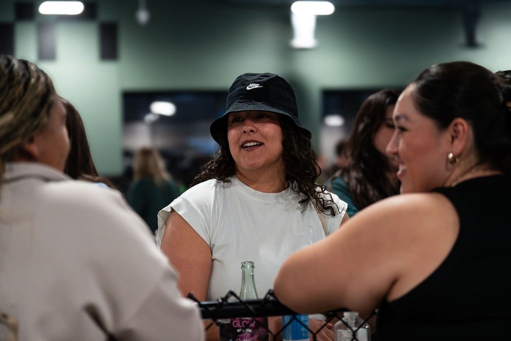 Three women are smiling and talking at a gathering or party, with a woman wearing a black Nike hat and white shirt in the center.