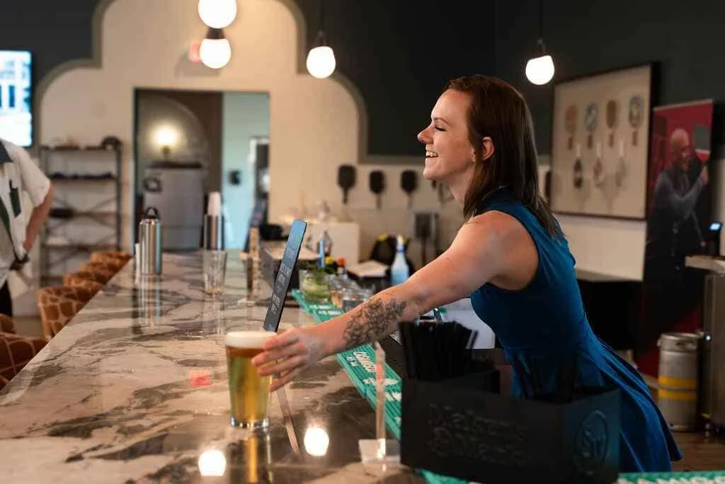 A bartender with tattoos, wearing a blue dress, smiling while serving a glass of beer at a bar with a marble countertop.