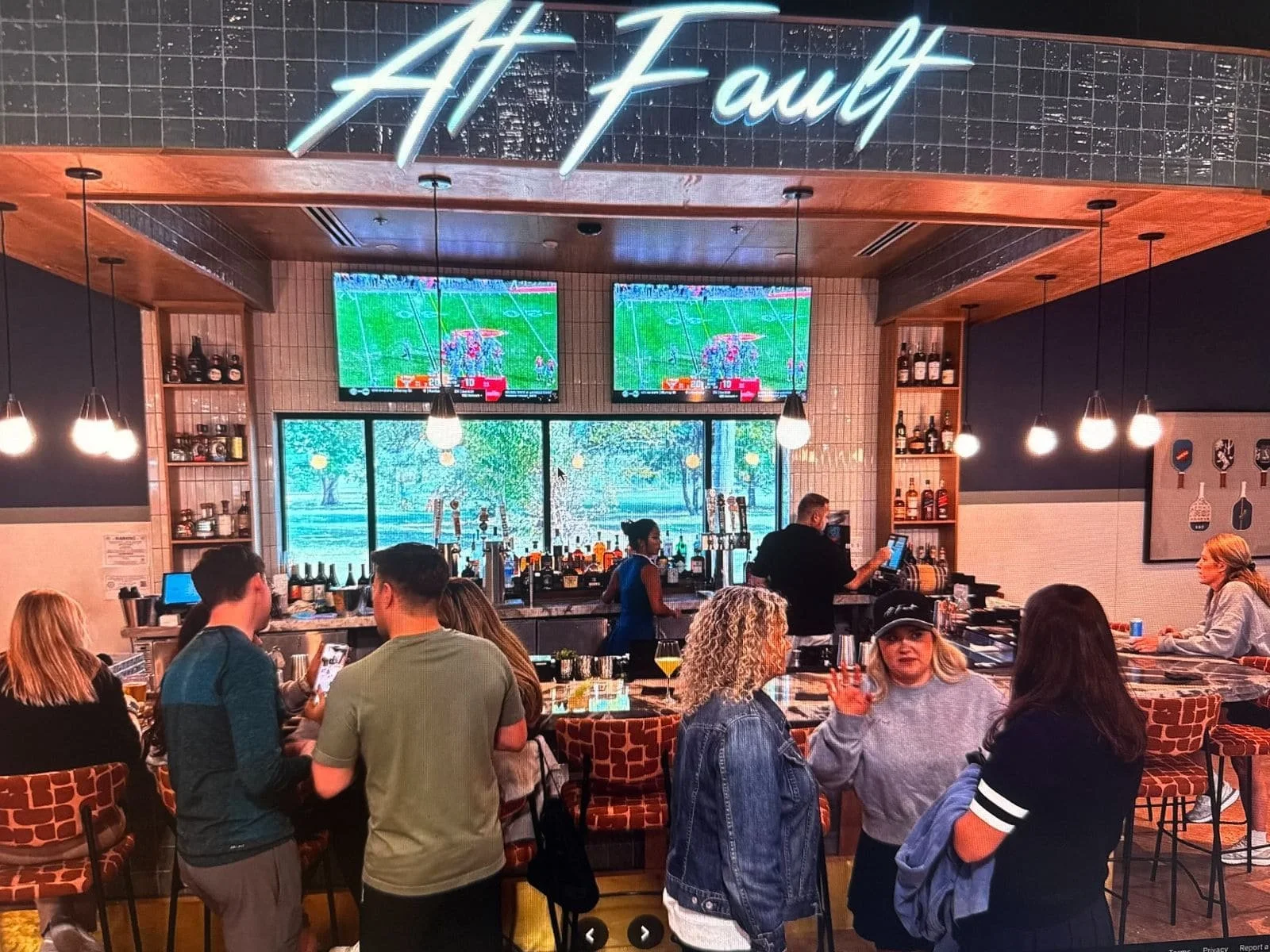 Interior of a bar or restaurant called 'At Fault' with people socializing and watching sports on multiple TVs. The bar area has shelves of bottles, and there are framed pictures of drinks on the wall. Pendant lights hang from the ceiling.
