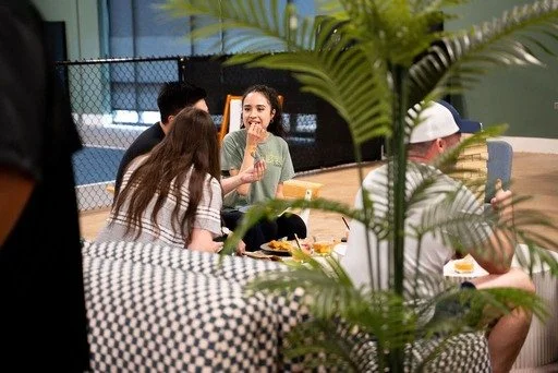 A group of people sitting around a table, socializing and eating in a casual indoor setting, with a large green plant partially obscuring the view.