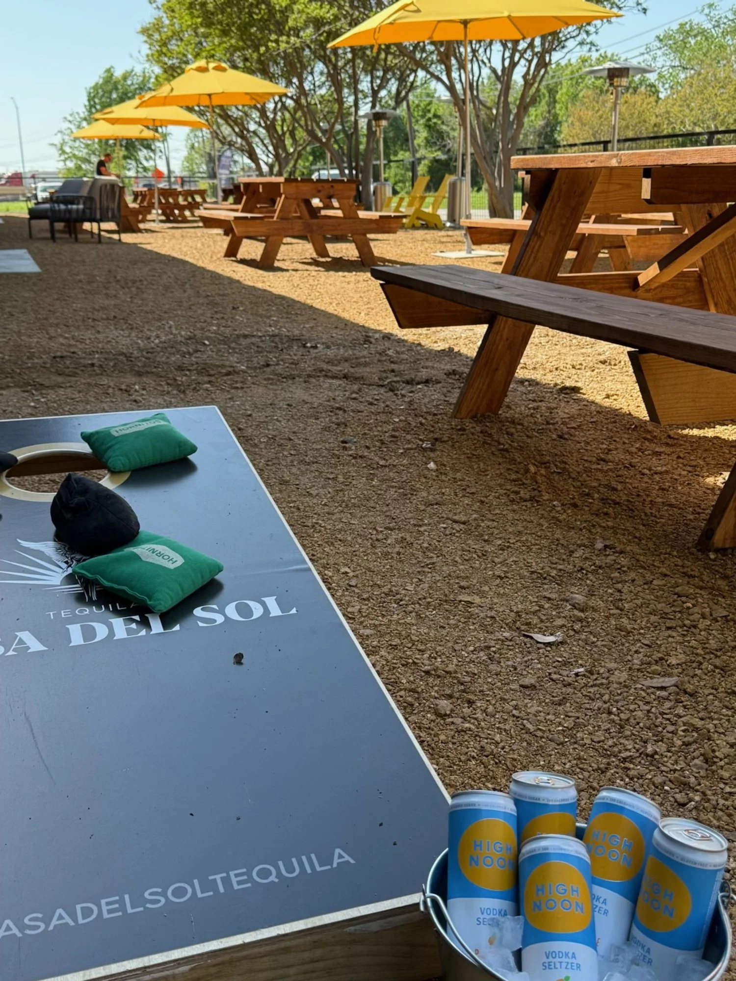 Outdoor seating area with wooden picnic tables, yellow umbrellas, and a table with cans of High Noon vodka seltzer, placed on a gravel surface with trees and a fence in the background.