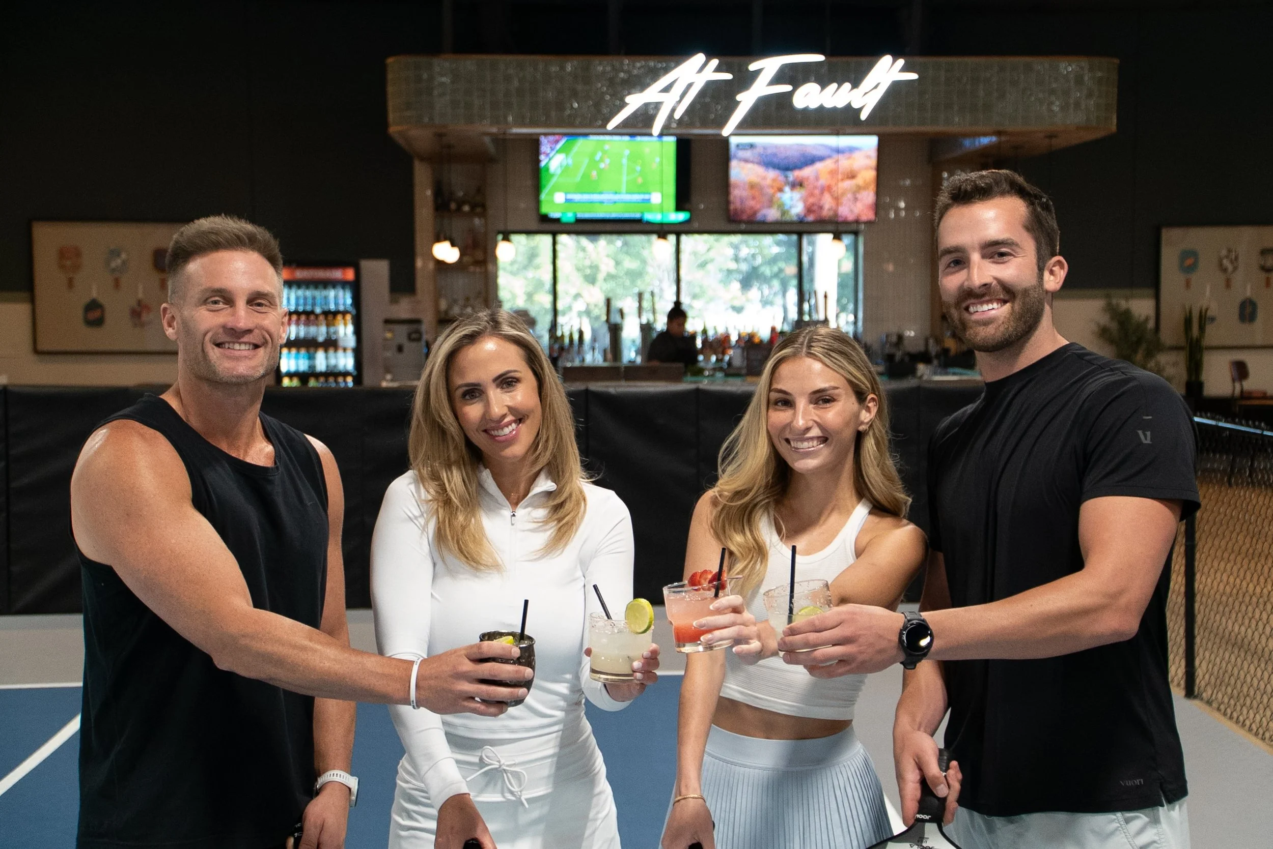 Four friends standing on tennis courts, holding cocktails, smiling, with a bar in the background and a sign that reads 'At Fawn' overhead.