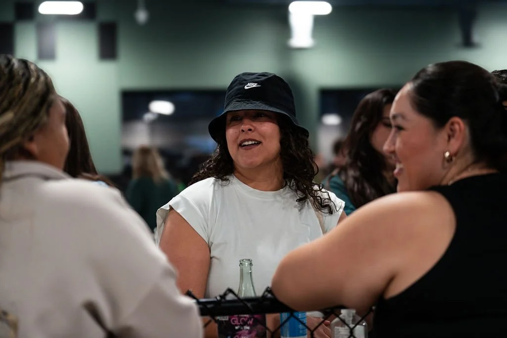 Three women in conversation, one wearing a black hat and white T-shirt, smiling and engaging in conversation at a social gathering.