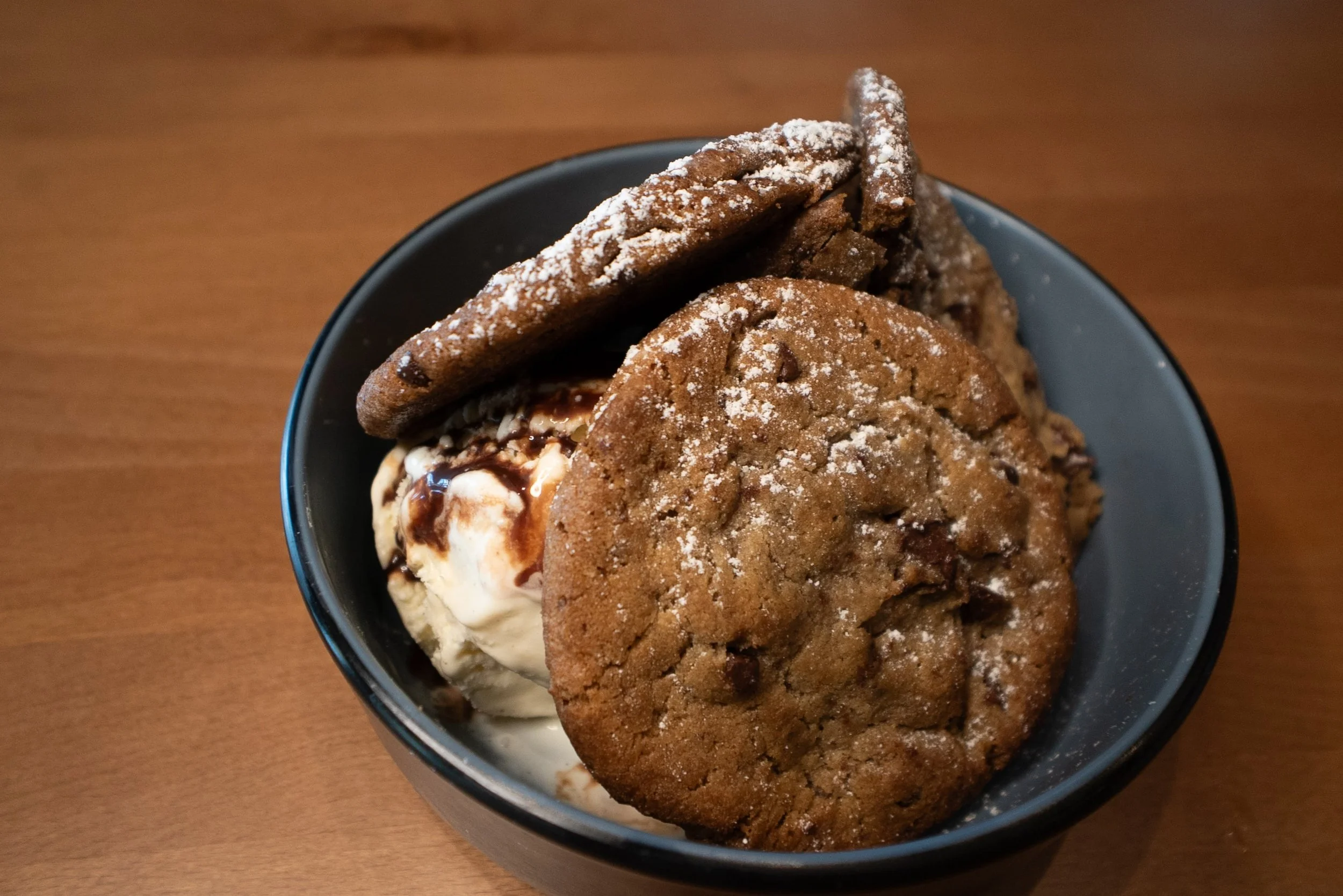 A bowl of vanilla ice cream topped with chocolate syrup, two large chocolate chip cookies, and two chocolate cookies dusted with powdered sugar.