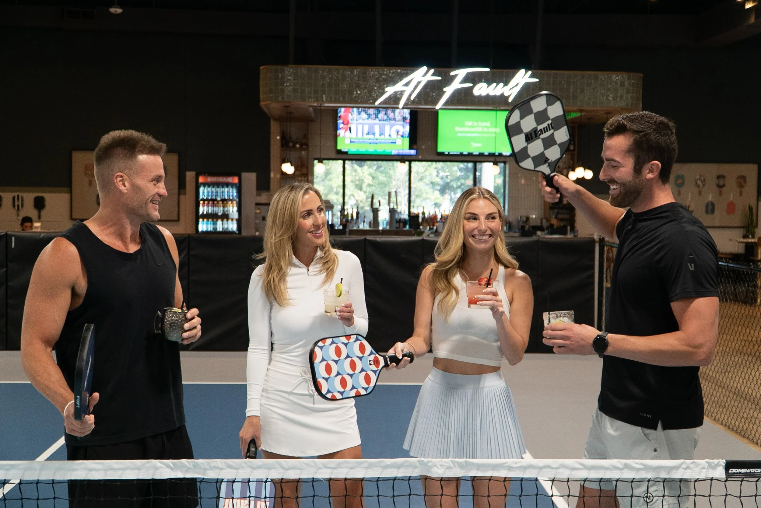 Four people standing on an indoor pickleball court, smiling and holding paddles and drinks, with a bar and TV screens in the background.