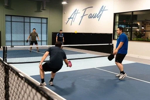 Four young men playing pickleball in an indoor court with a sign that says 'A Fault' on the wall.