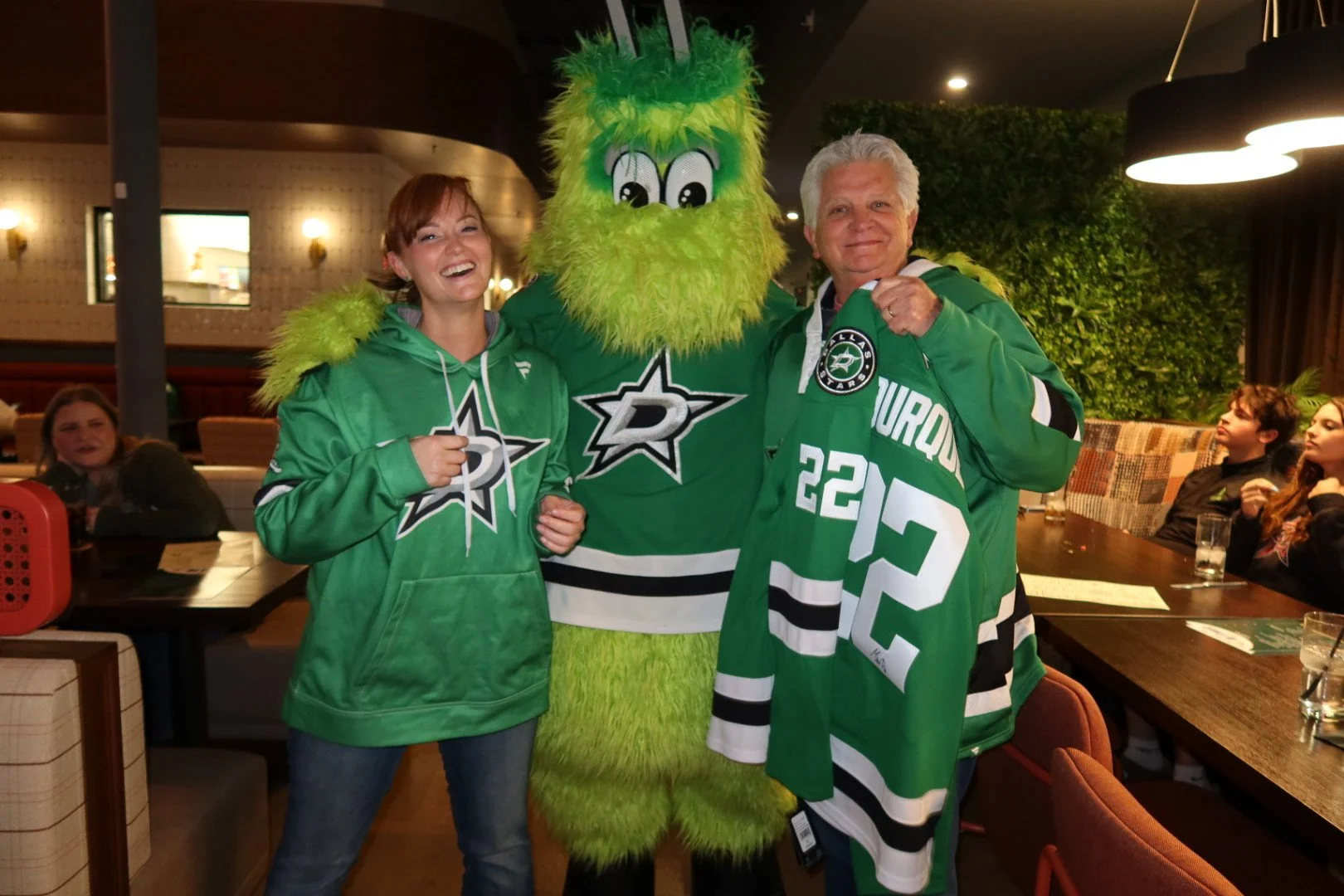 A woman, a person in a Dallas Stars mascot costume, and a man in Dallas Stars hockey jerseys posing together inside a restaurant or bar, smiling and showing team spirit.