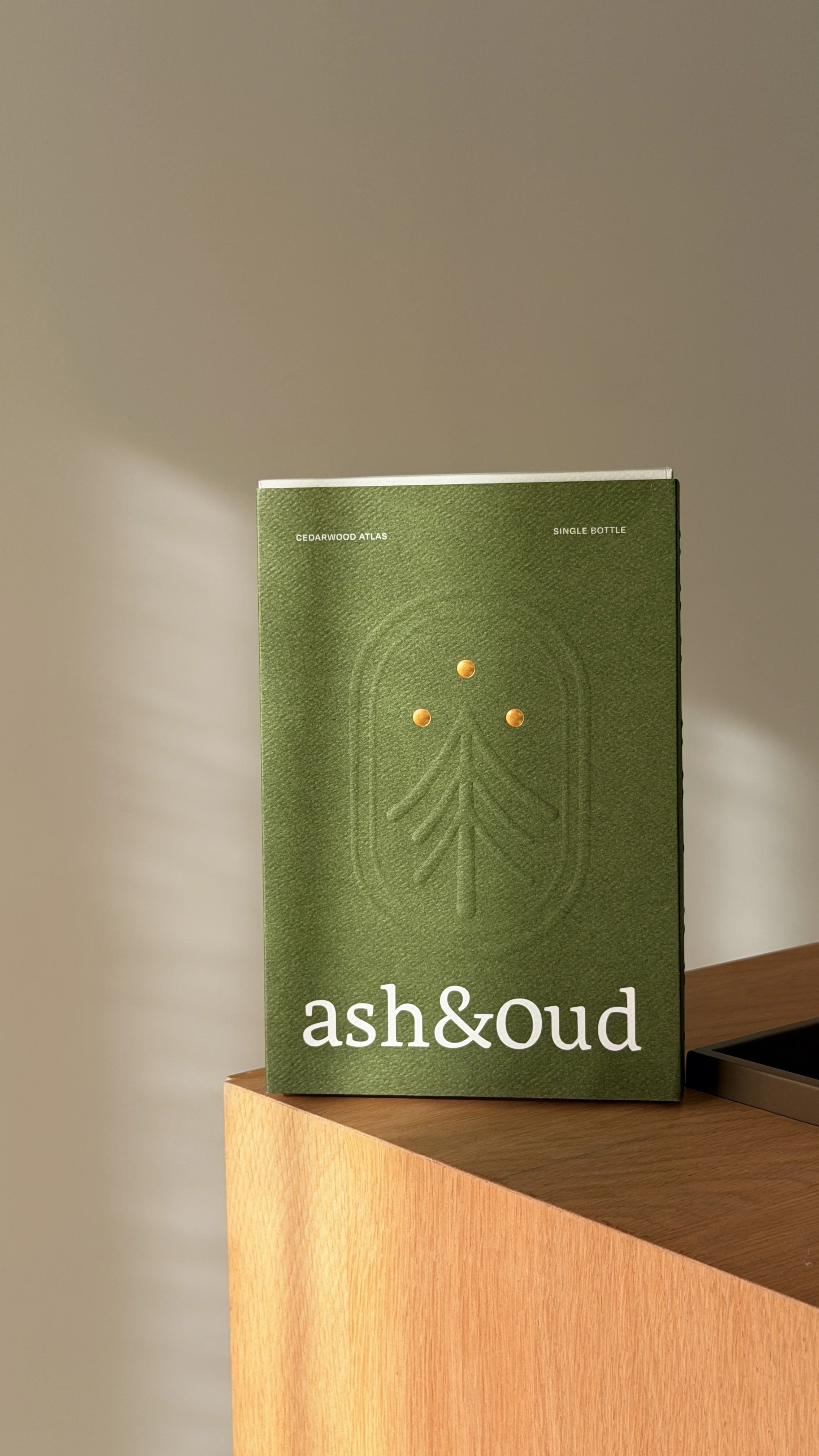 A green box labeled "ash&oud" with an embossed tree design and three yellow screws on the front, placed on a wooden surface.