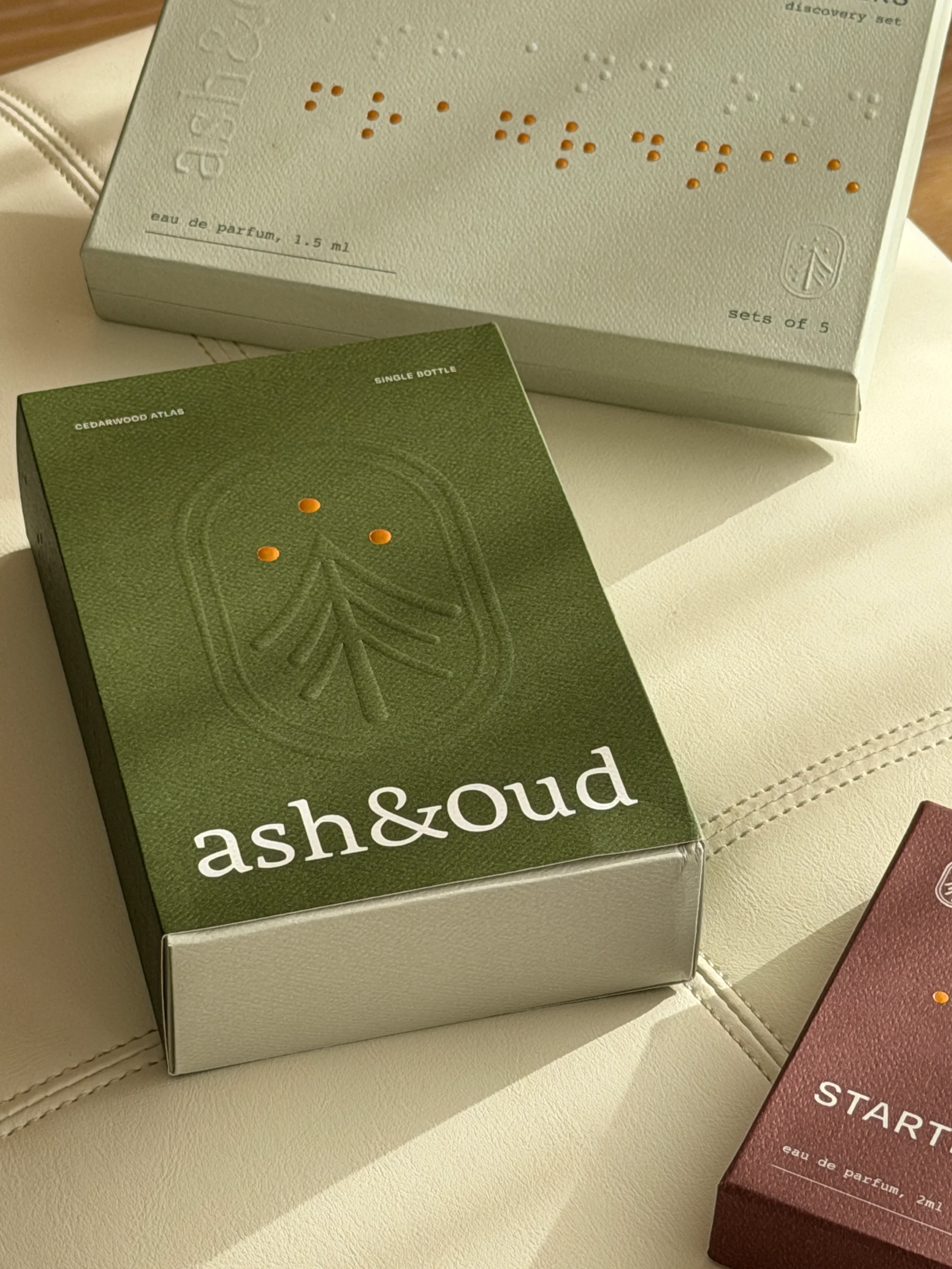 Three sets of perfume boxes with minimalist design, one labeled ash&oud in green, another gray with Braille and embossed design, and a smaller reddish-brown box labeled START in white, placed on a cream-colored surface.