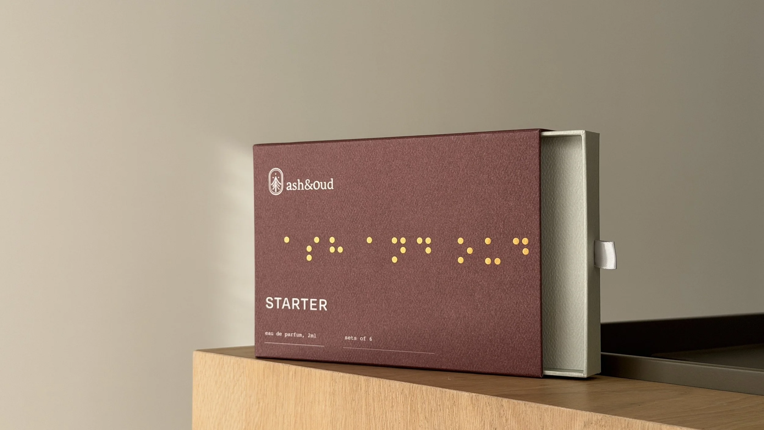 A box of ash & oud perfume starter set with six 2ml eau de parfum bottles, placed on a wooden surface against a plain wall.