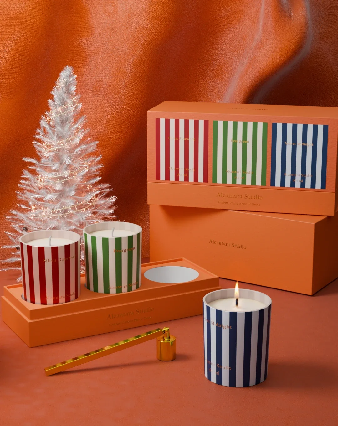A white decorated Christmas tree with lights next to three striped candles in red, green, and blue, placed in an orange box with matching packaging in the background.