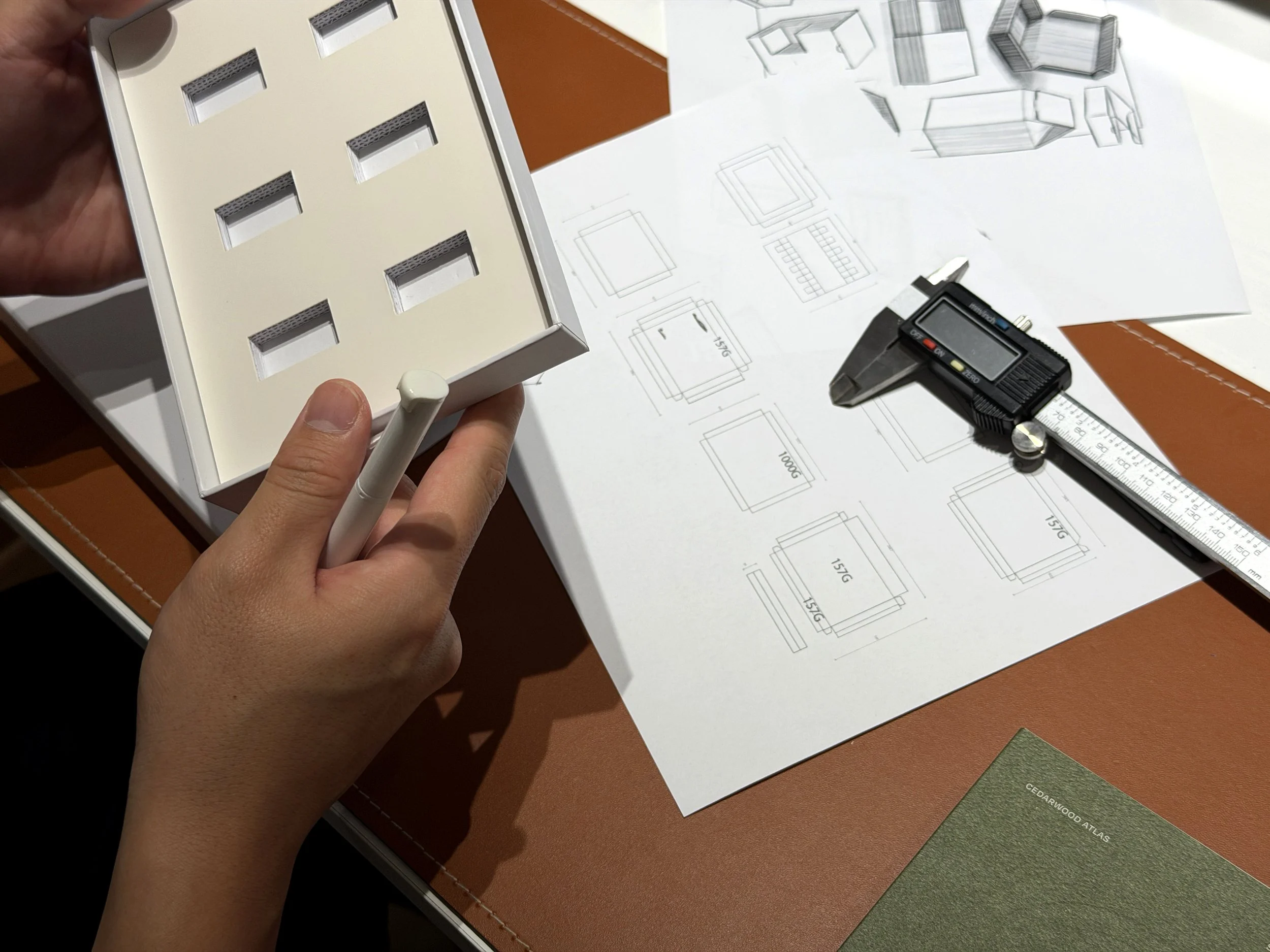 Person holding a white box with six rectangular compartments. On the table are technical drawings, a digital caliper, and a green sample card labeled 'CEDARWOOD ATLAS'.