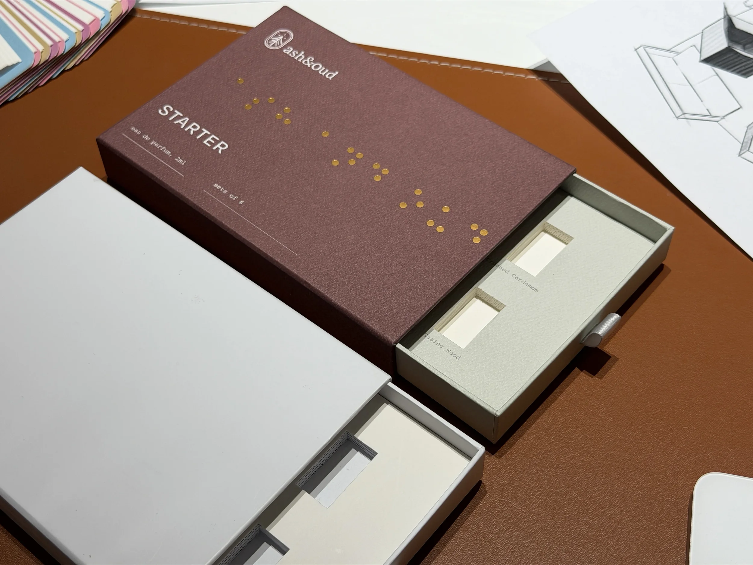 Open sample perfume discovery set reveals two small perfume bottles labeled 'Santal Wood' and 'Unscented Cardamom' inside a white box with a textured surface, placed on a brown desk near a piece of paper with architectural drawings.