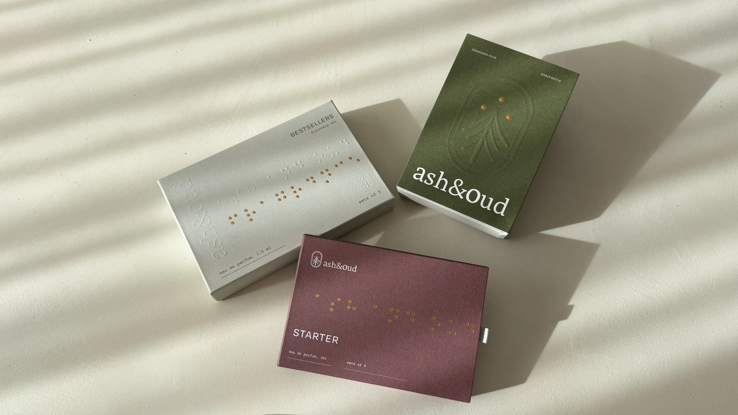 Three boxes with minimalistic design, one green, one white, and one pink, labeled as ash&oud, placed on a white surface with sunlight and shadow.