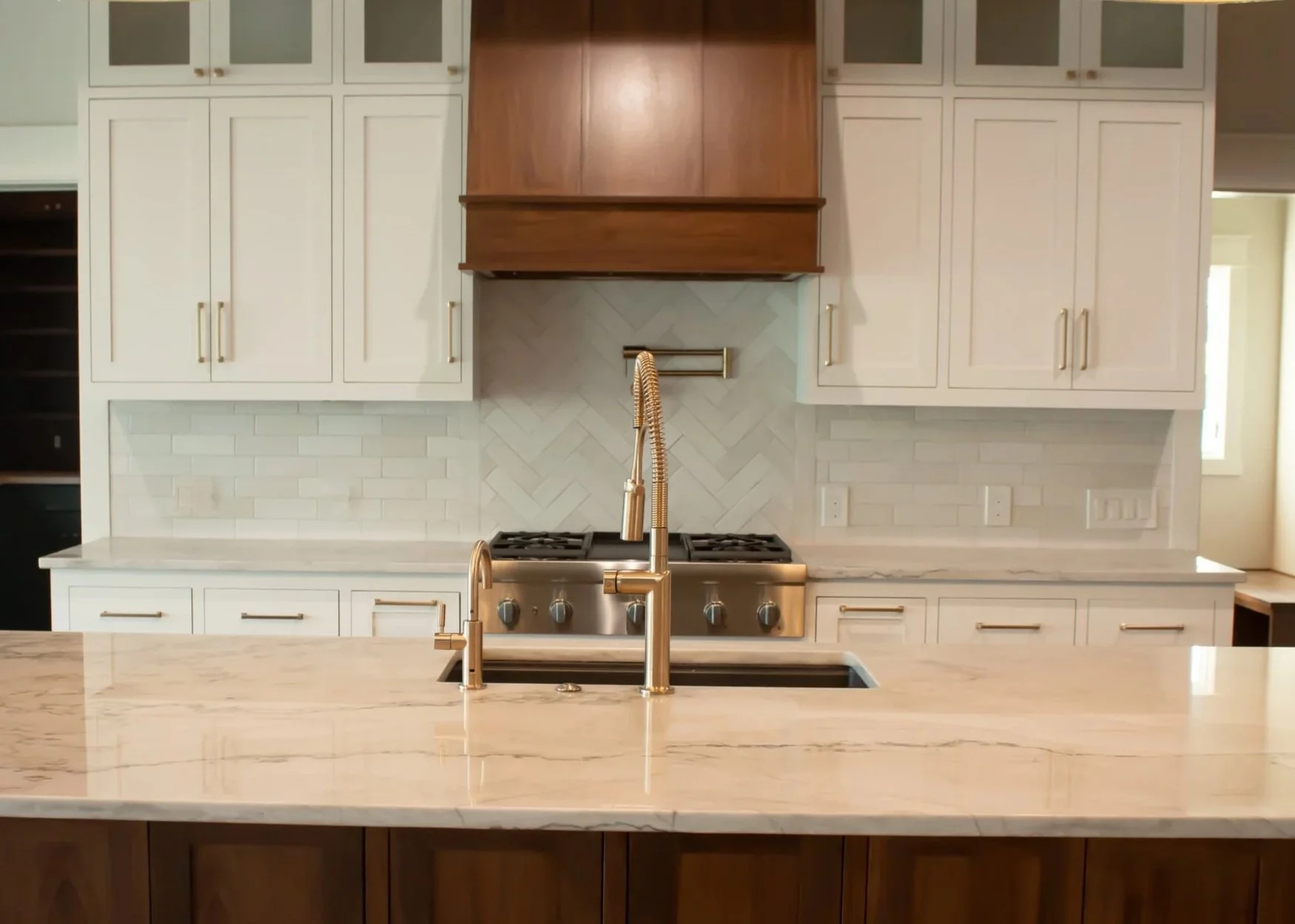 Modern kitchen with white cabinets, a marble countertop, and a stainless steel stove with three burners. Brass fixtures on the sink and a wooden range hood above the stove. The backsplash is white herringbone tile.