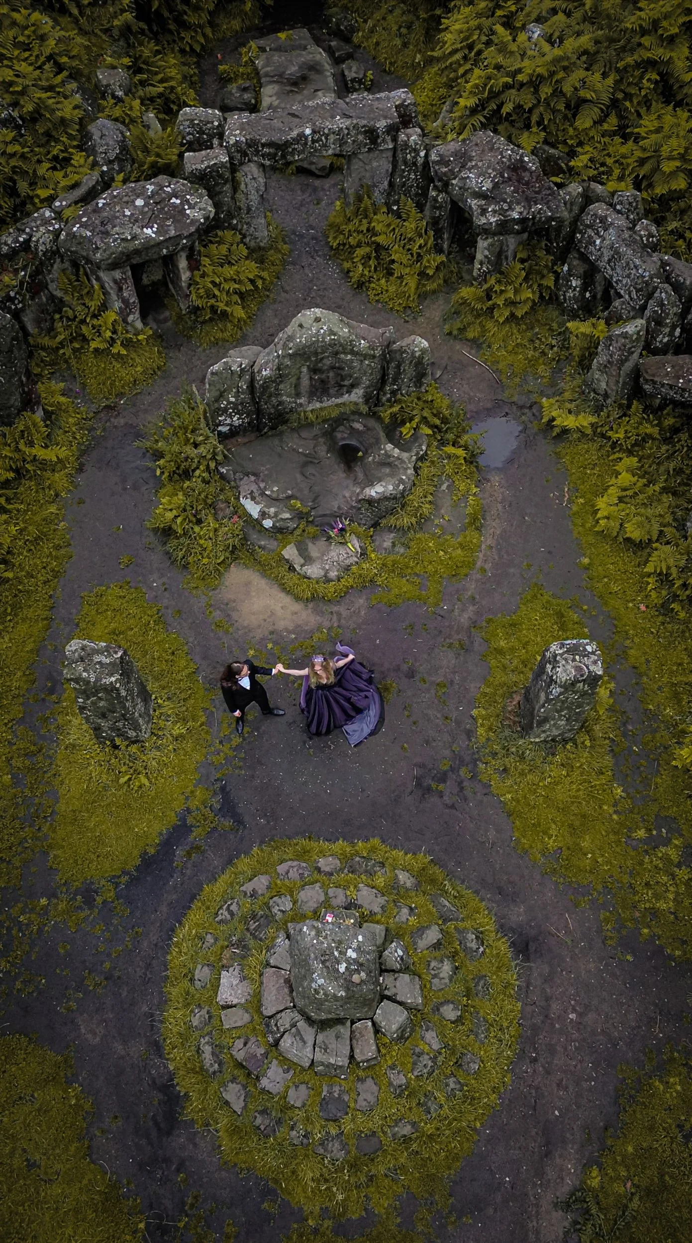 An aerial view of two people in formal attire, dancing and holding hands in a garden with large rocks, moss, and ferns.