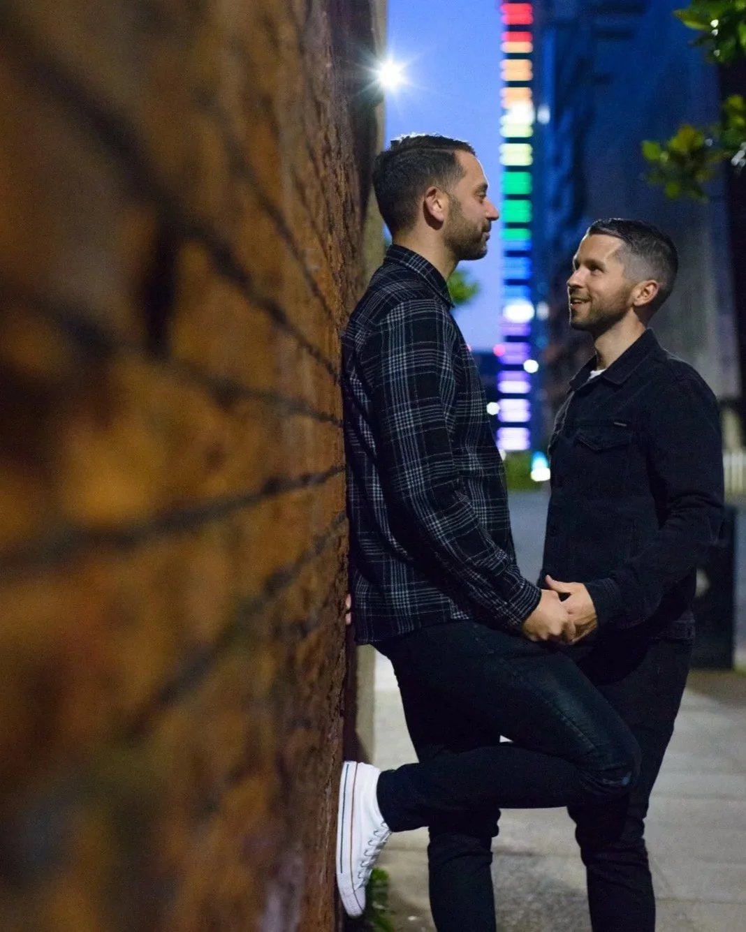 A couple holding hands and smiling at each other on a city street at night, with colorful lights in the background.