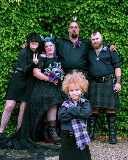 Group of five people standing outdoors in front of green hedges, with a woman in the center holding a bouquet of flowers, and a child with curly hair standing in front of them.