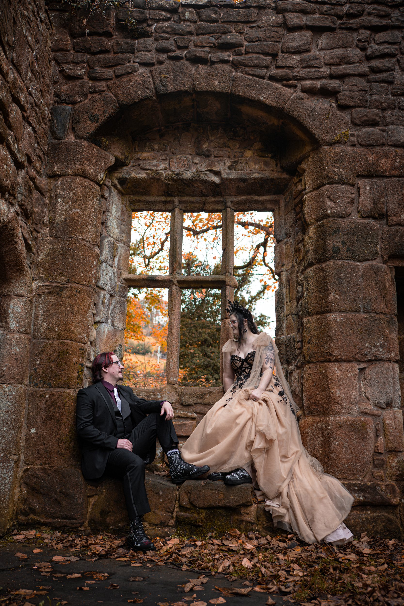 A Husband and Wife are sitting in a window space of a castle ruin. Both wearing black boots with white spiderweb designs. Staring into each other's eyes lovingly. Post-wedding shoot.