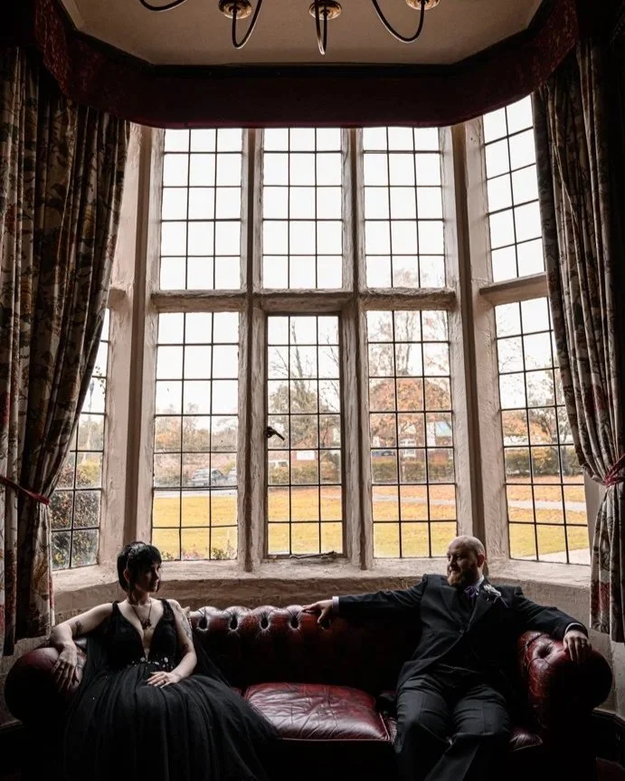 Two people sitting on a red leather couch beneath a large bay window with multiple panes, flanked by patterned curtains, in a room with a chandelier on the ceiling, during daytime.