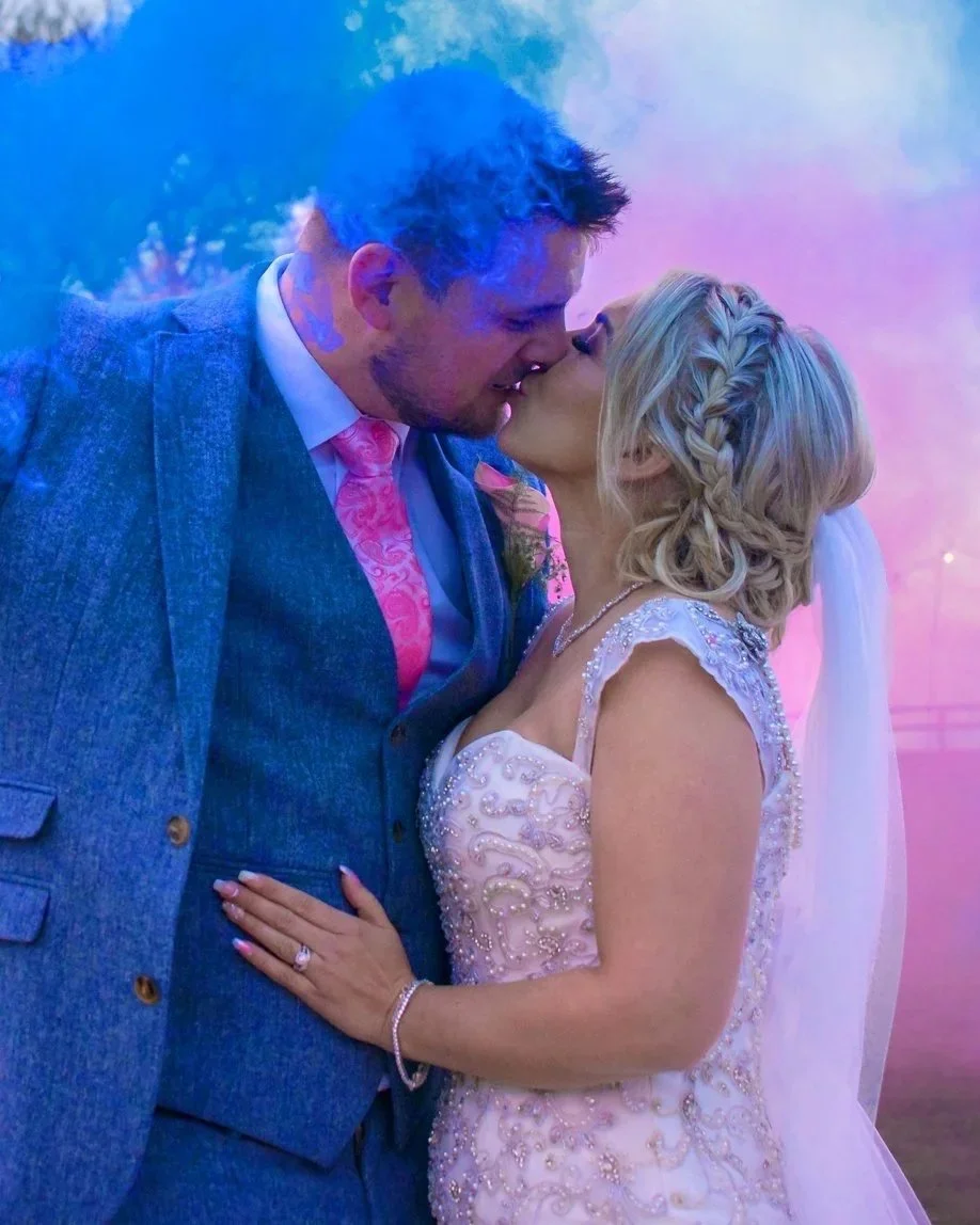 A newlywed couple sharing a kiss at their wedding reception, with colorful smoke in the background.
