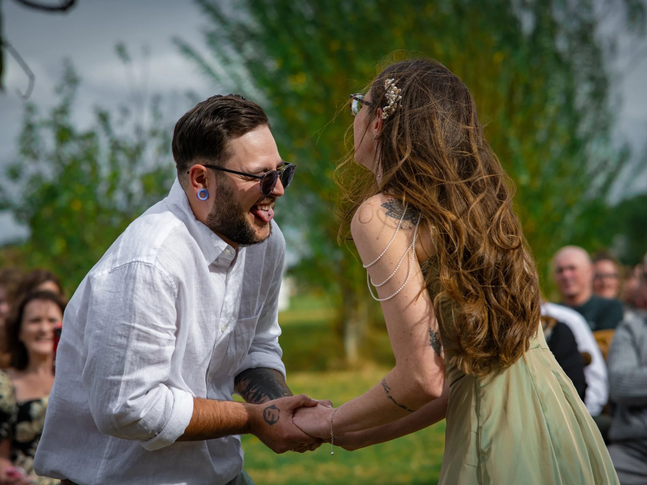 A man with sunglasses and gauges holding hands and smiling with a woman with long curly hair and decorated hair accessories at an outdoor event.