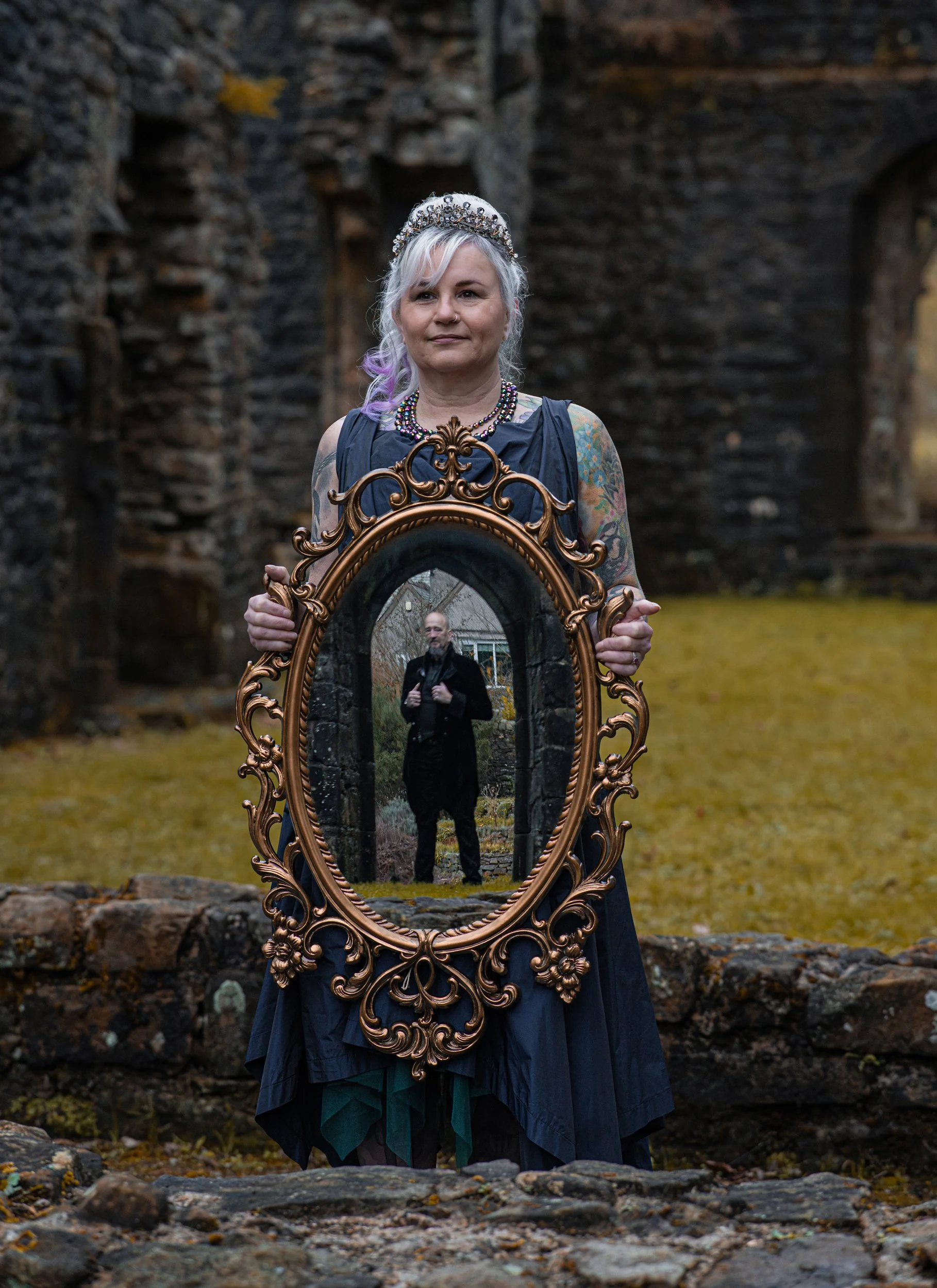 A woman holding a mirror and in the reflection you can see her husband. Standing within a gothic arch doorway, holding his jacket collar in both hands. Gothic post wedding shoot.