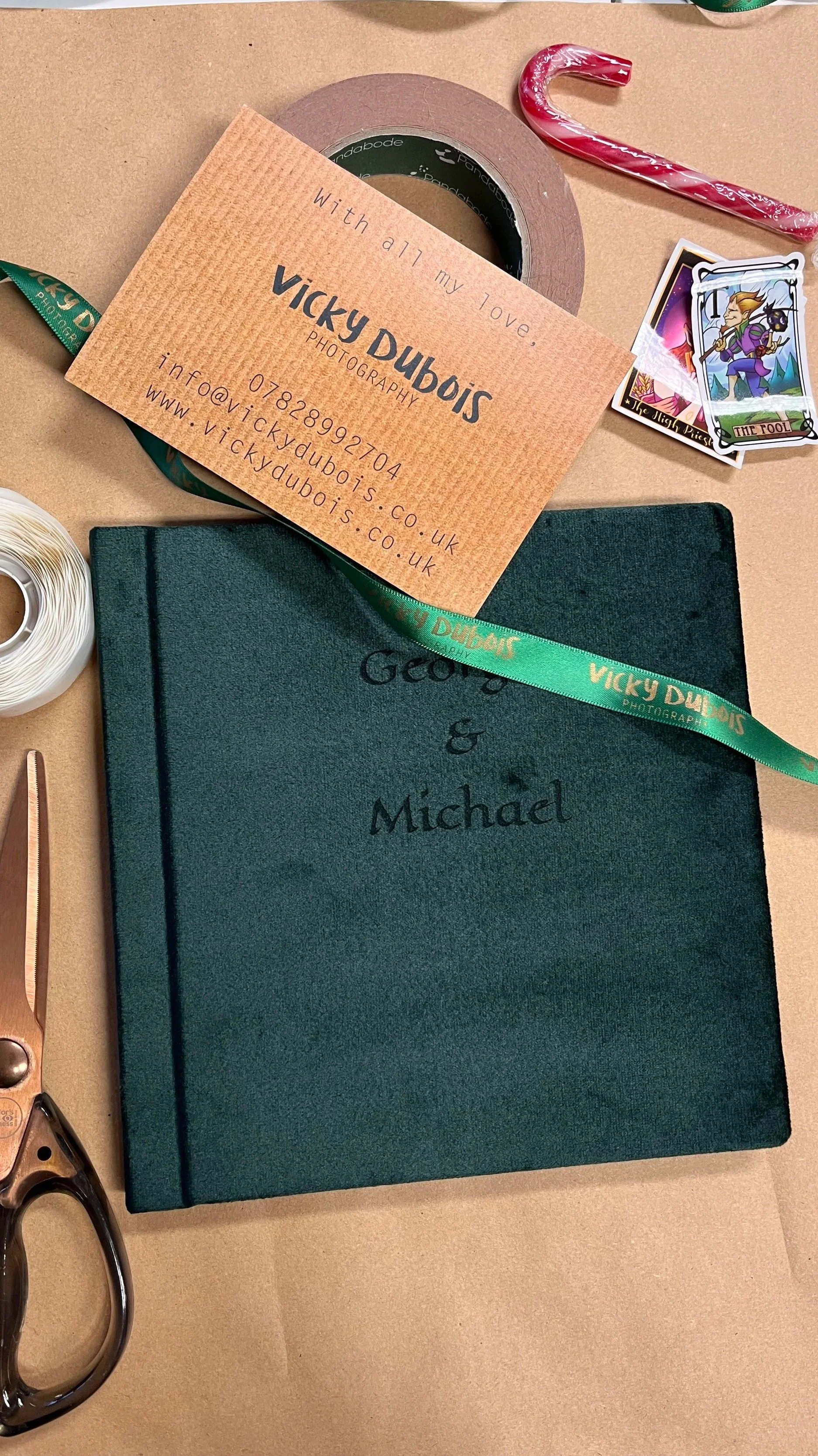 A teal photo album with the names 'George & Michael' embossed on the cover, a Cricut cutting tool, a roll of tape, a red and white striped candy cane, a business card for Vicky Dubois Photography, and a sticker of a cartoon character from 'The Night Prism'.