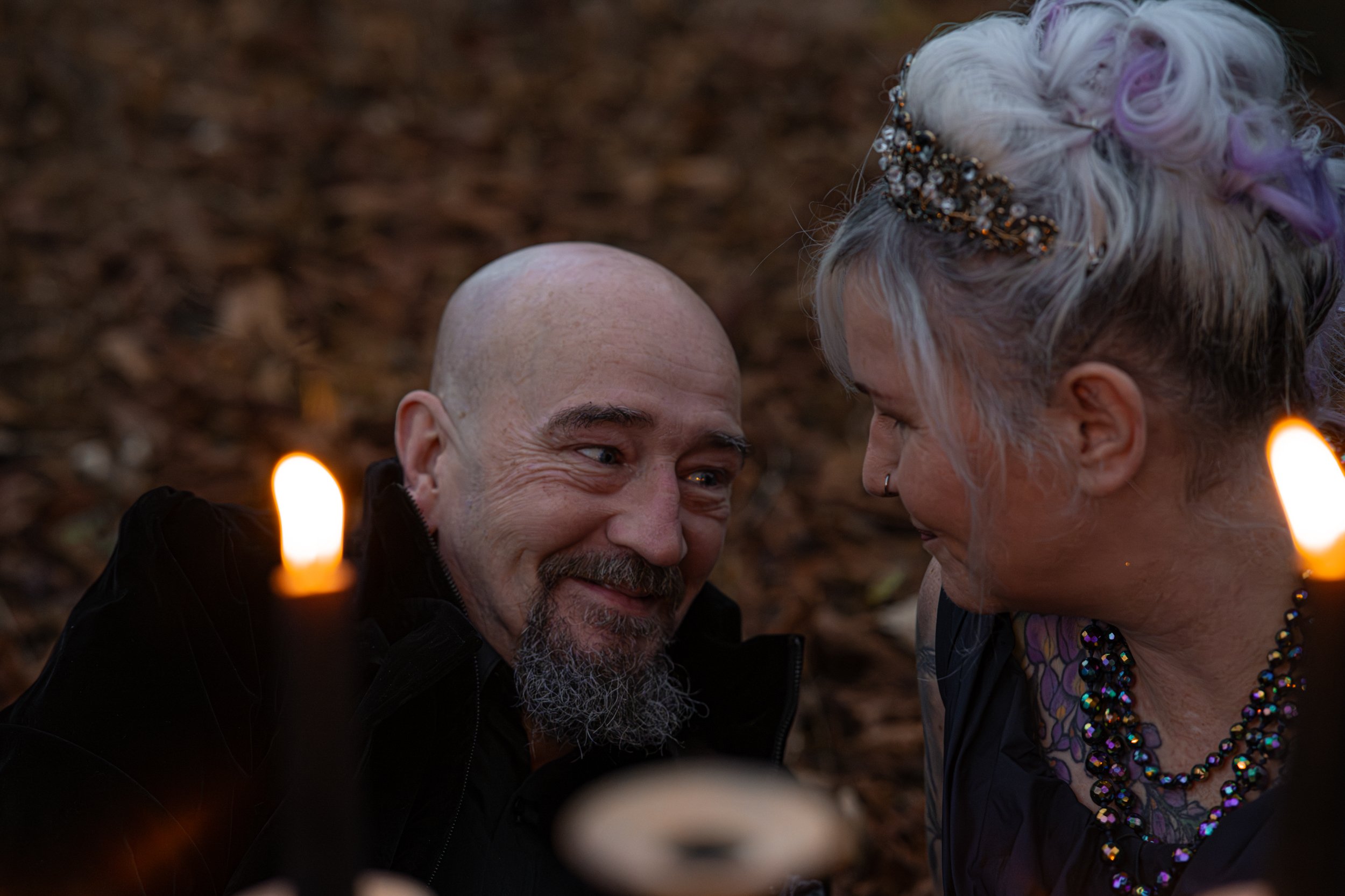 An alternative couple staring into each other's eyes lovingly with smiles on their faces. The husband's face is the centre face with the side of the wife's face to the right. A candelabra is out of focus in the foreground.