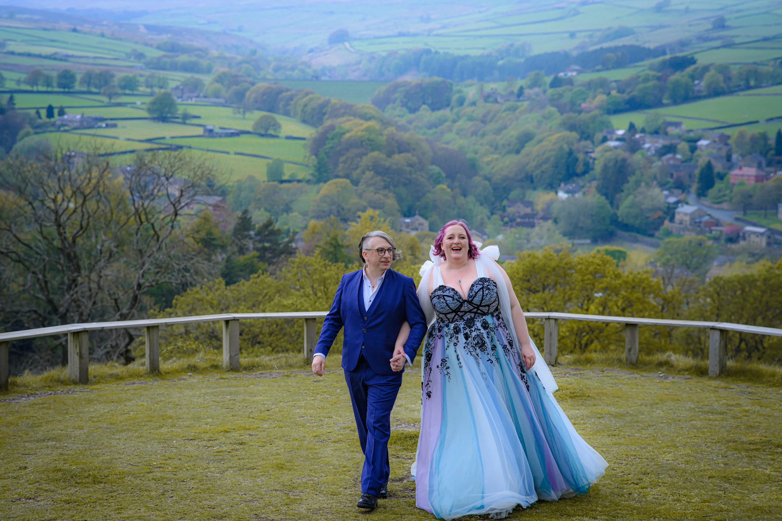 Two women holding hands and smiling, walking outdoors on a grassy area with a scenic landscape of rolling hills and trees in the background. One woman wears a blue suit, and the other wears a colorful, flowing gown.
