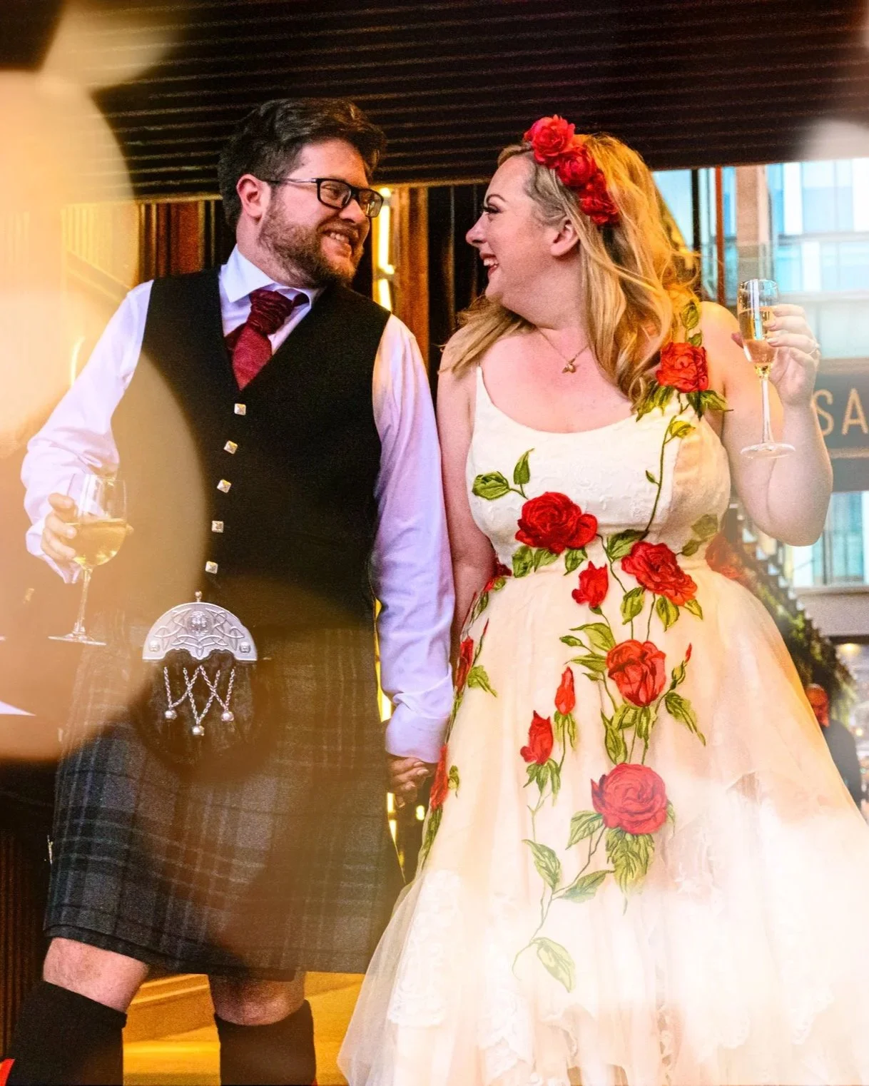 A man and woman dressed in wedding attire are celebrating, holding glasses of champagne and smiling at each other, in an indoor setting with warm lighting.