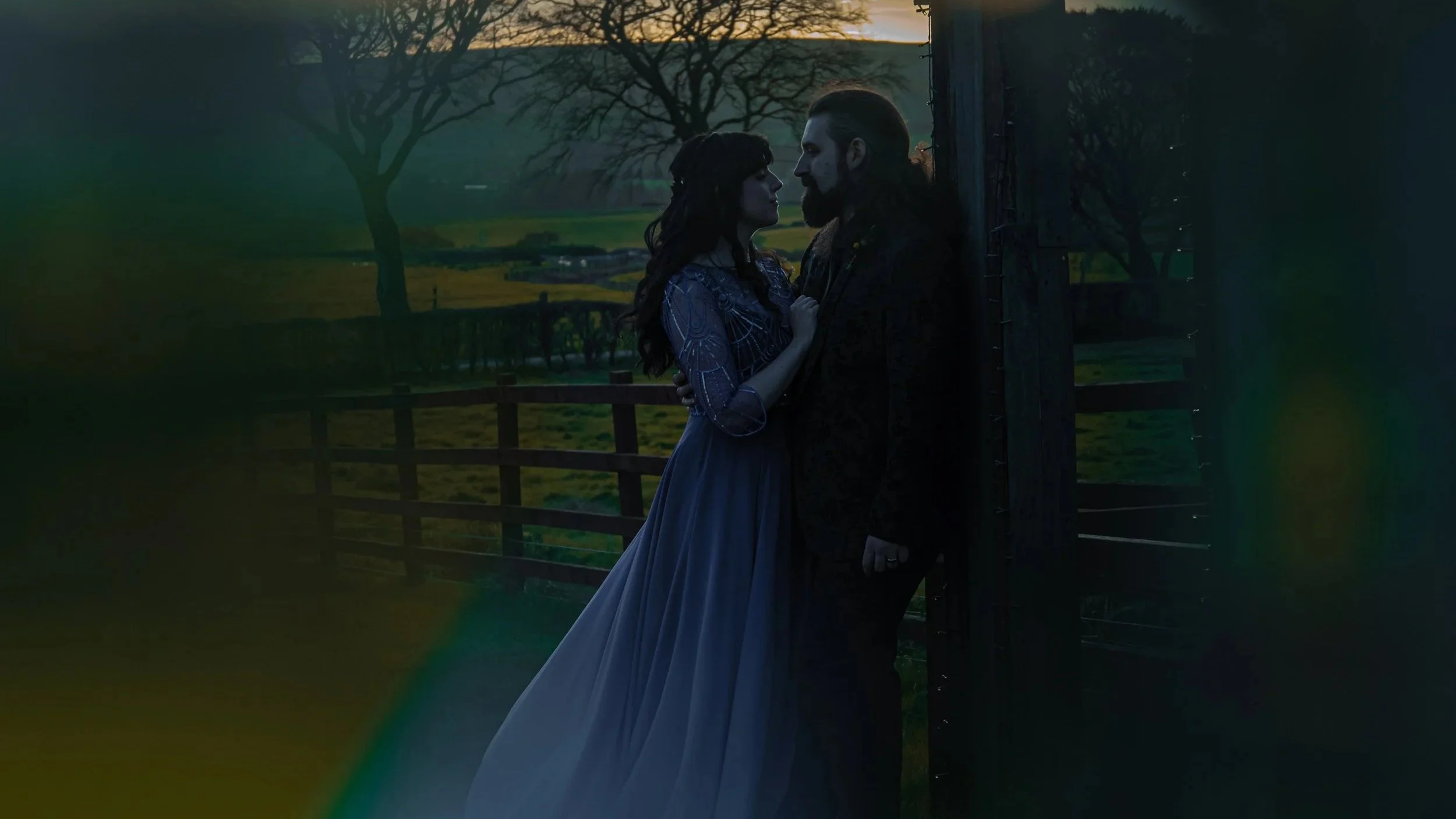 A man and woman stand close together, gazing into each other's eyes near a dark wooden fence in a rural setting at sunset, with leafless trees and open fields in the background.