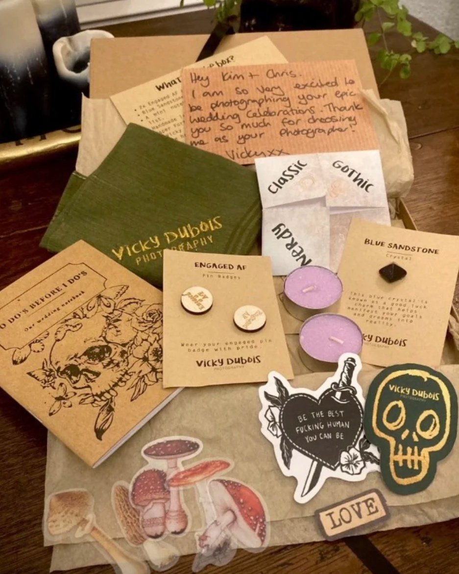 A collection of items including handwritten notes, stickers, candles, pins, and decorative paper on a wooden surface. The notes contain personalized messages, and the stickers feature skulls, a heart with an inspirational quote, and mushrooms.