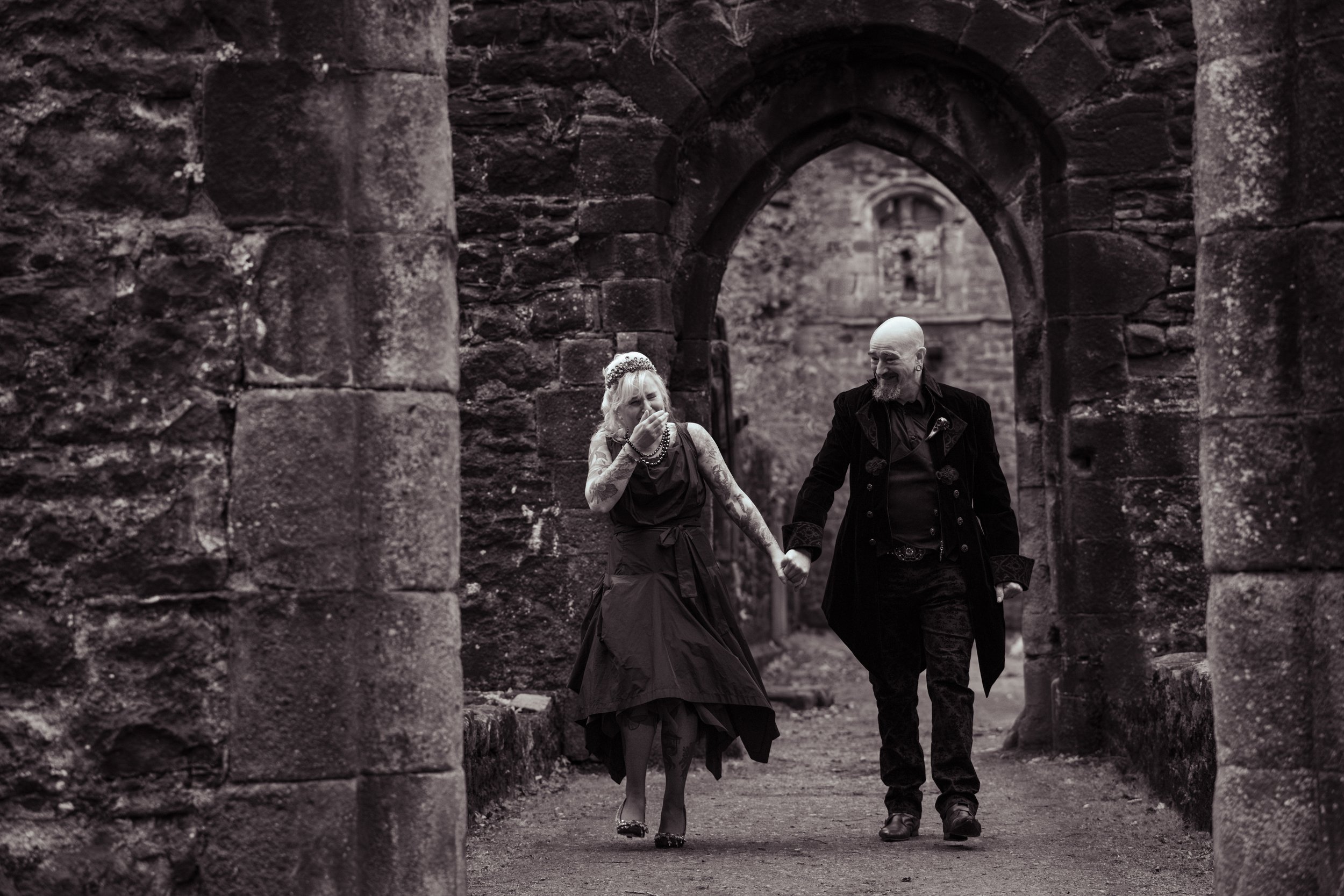An alternative Husband and wife walking hand in hand through castle ruins. Both laughing. Post-wedding shoot.