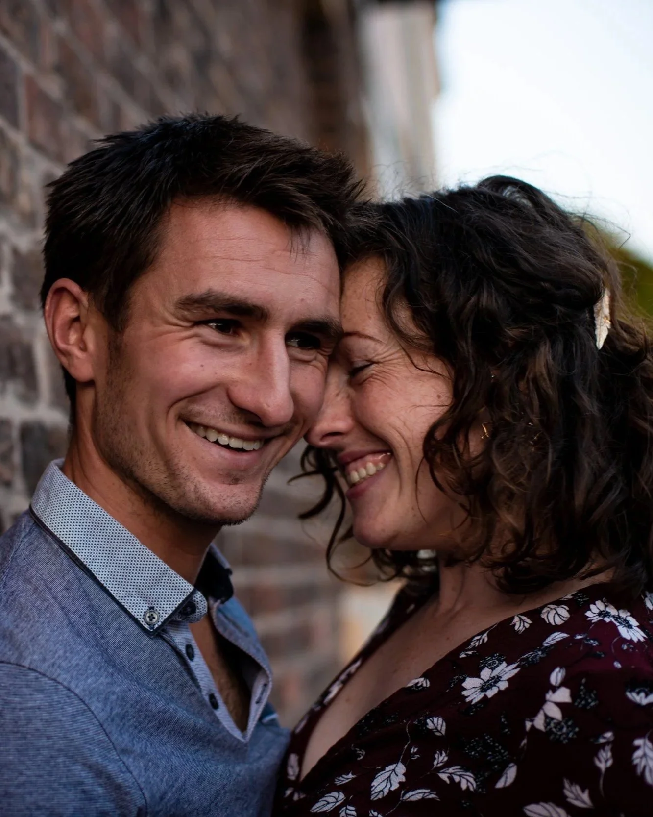 A smiling man and woman in a close embrace, touching foreheads and eyes closed, standing outdoors against a brick wall.
