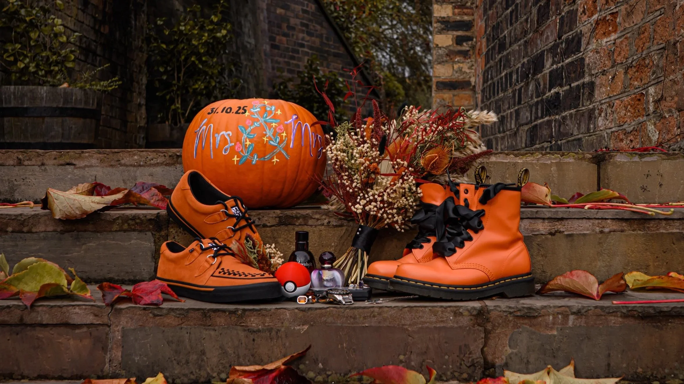 Memorial setup on steps with orange sneakers, a pumpkin decorated with 'Mrs. & Mrs.', flowers, fallen leaves, and small memorabilia items.