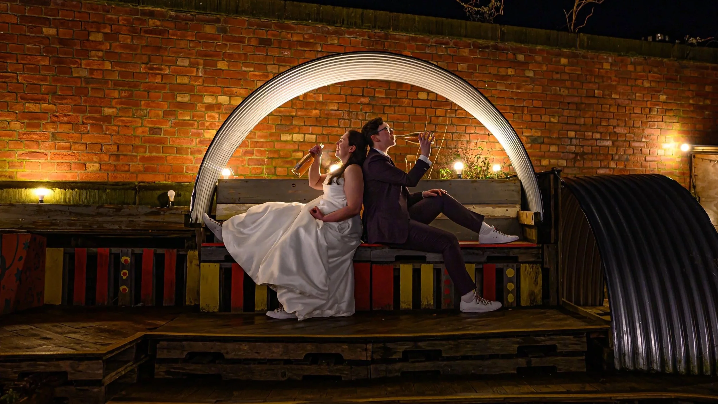 Lesbian couple on their wedding day drinking from champagne bottle in a reclamation style garden at Grub in Manchester. The couple are sat back to back in a suit and dress just chilling and drinking.