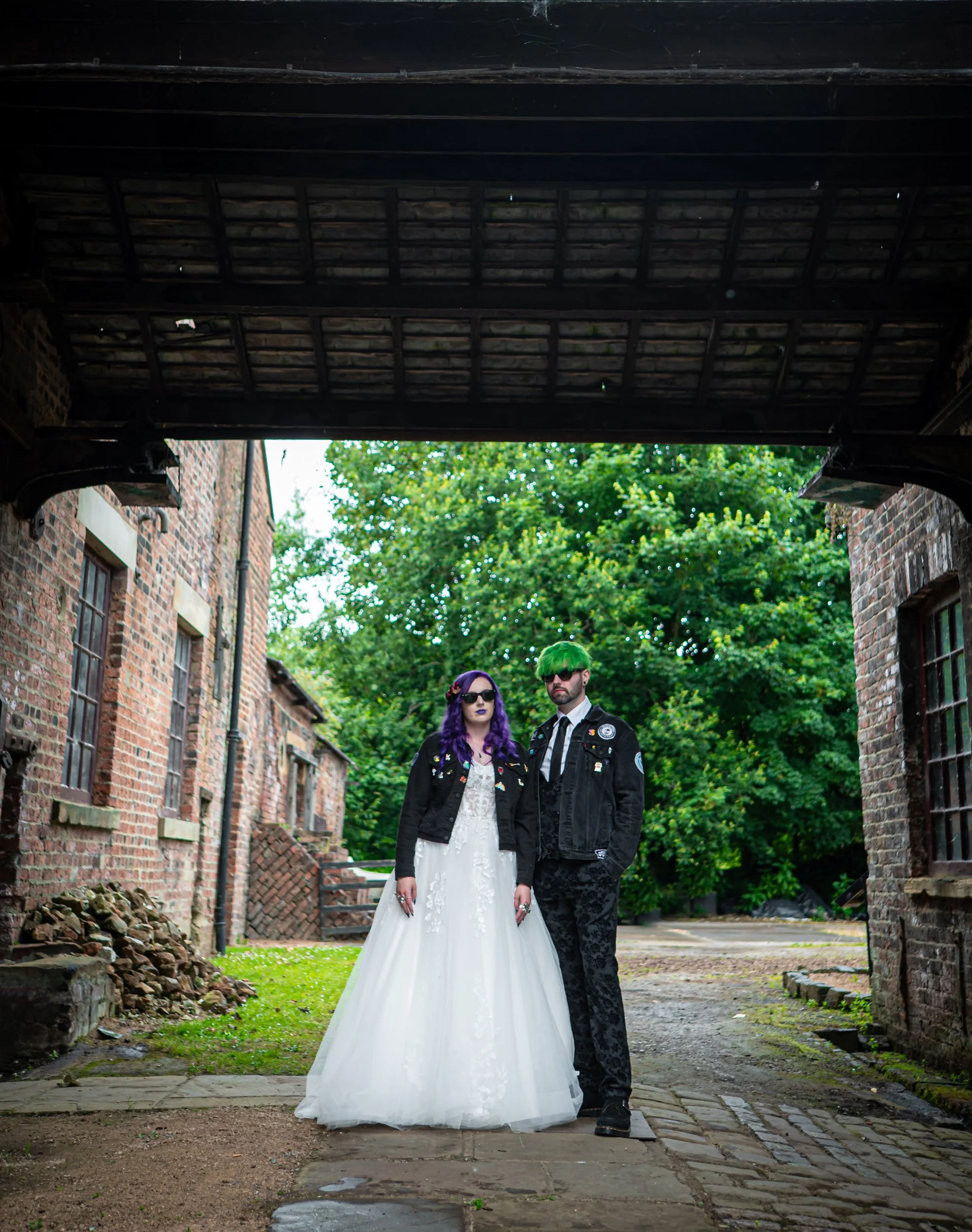 A couple with alternative styles standing together outdoors in front of an alleyway. The woman has purple hair and wears a white dress with a black jacket, while the man has green hair and wears sunglasses, a black jacket, and camouflage pants. The background features old brick buildings and green trees.