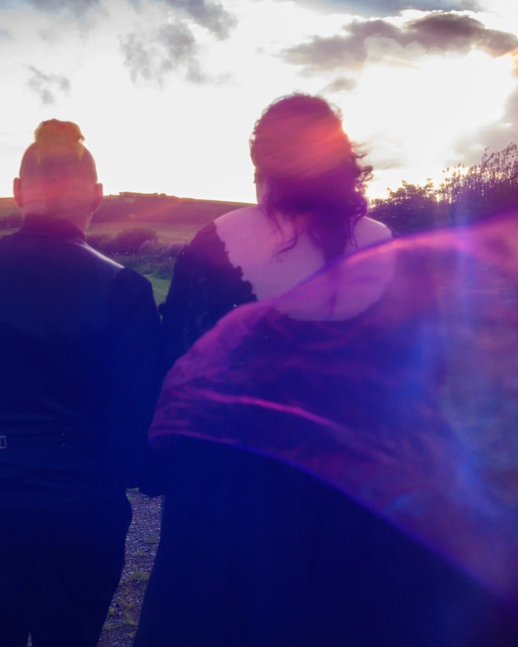 Two silhouetted people standing outdoors, with a sunset or sunrise sky and clouds in the background, and lens flare effects.