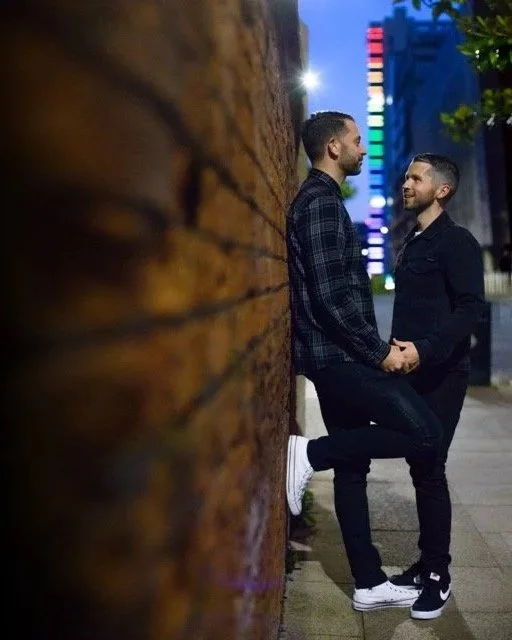 Two men are standing on a city sidewalk, facing each other and holding hands. They are near a brick wall, with colorful city lights in the background, during evening or night.