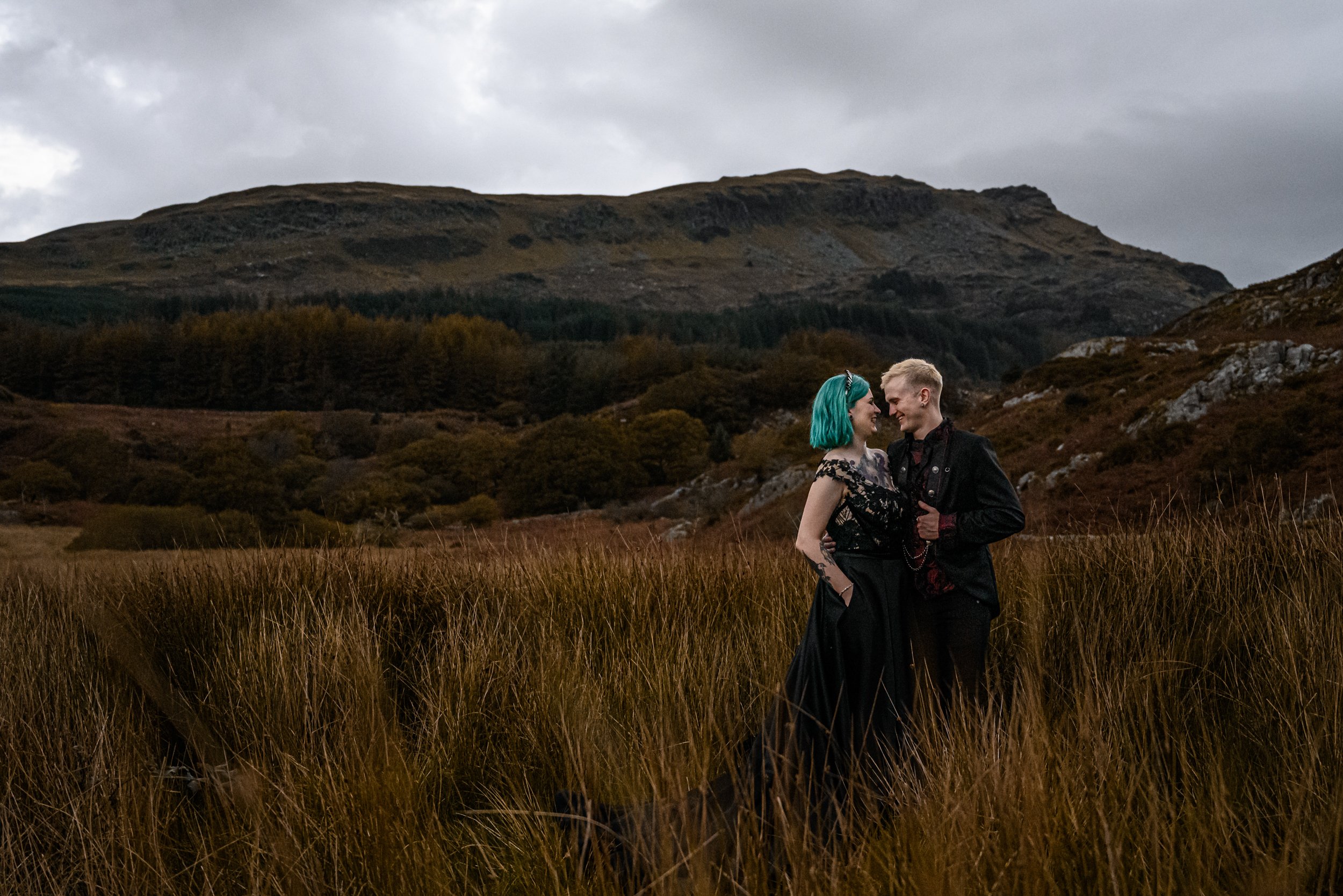 Tattooed, alternative couple on their wedding day, romantically stood in a wild field in North Wales. Dramatic black, gothic dress and suit. Bride has green hair and groom has metal pocket chain
