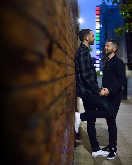 Two men are standing outside at night, holding hands, and looking into each other's eyes in front of a city street scene with colorful lights.