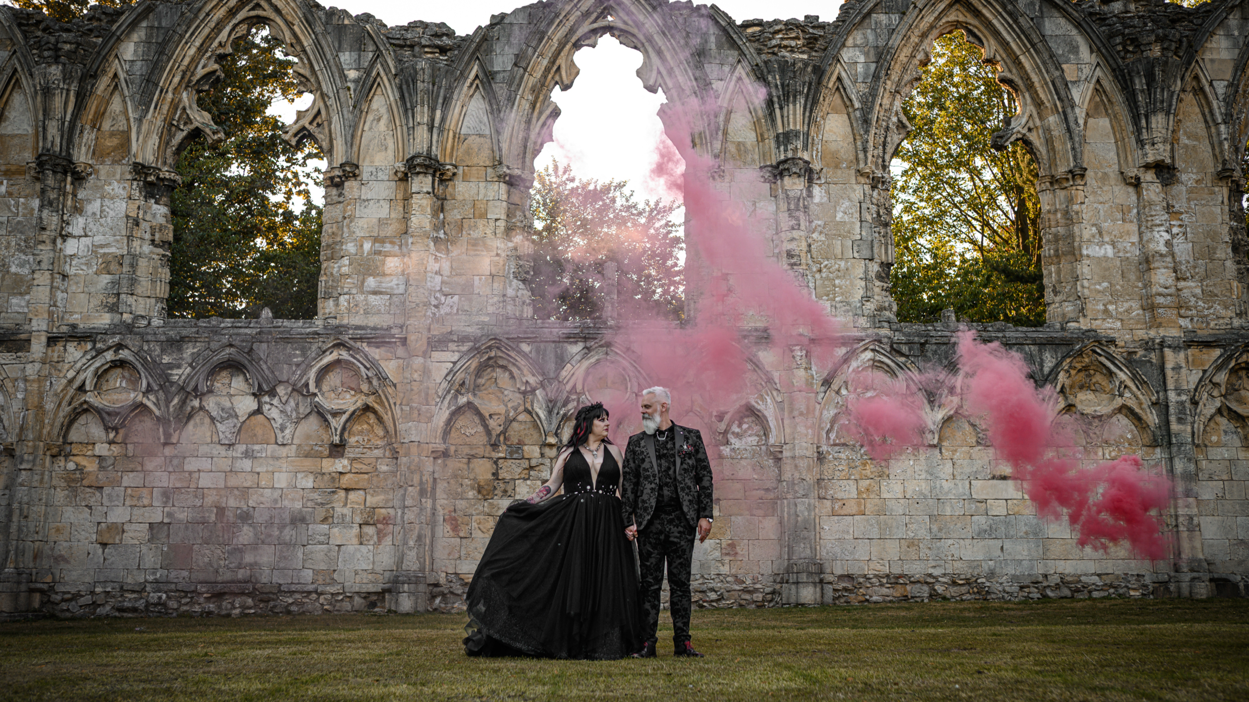 A couple dressed in gothic clothing stands hand-in-hand in front of a ruined stone building with arched windows. Pink smoke billows around them during daytime.