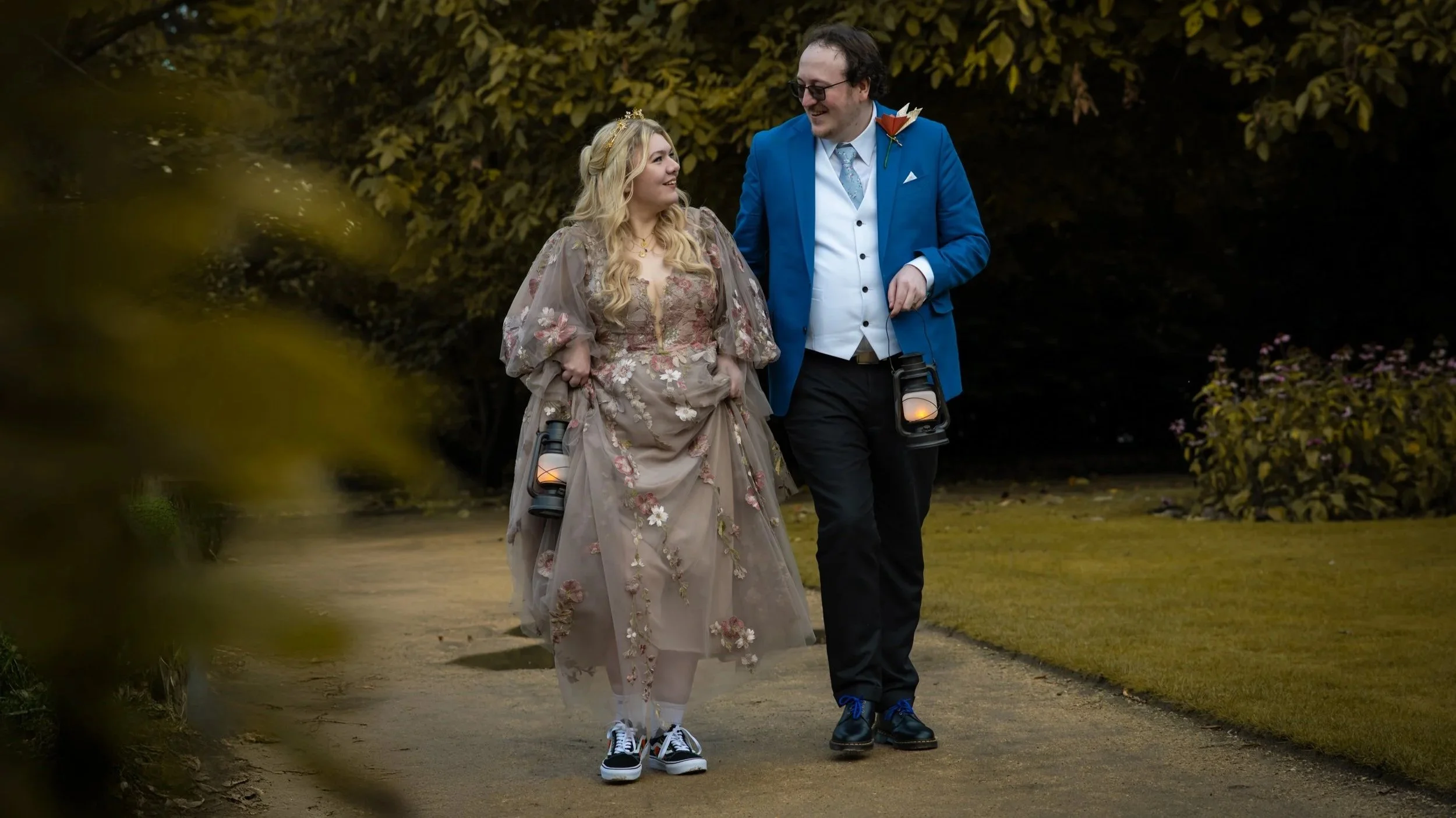 A couple walks outdoors on a dirt path surrounded by greenery. The woman wears a detailed, floral-patterned, vintage-style dress with sheer sleeves, and the man wears a bright blue suit jacket with a white shirt, black pants, and sneakers. They look at each other and smile, each carrying a lantern.