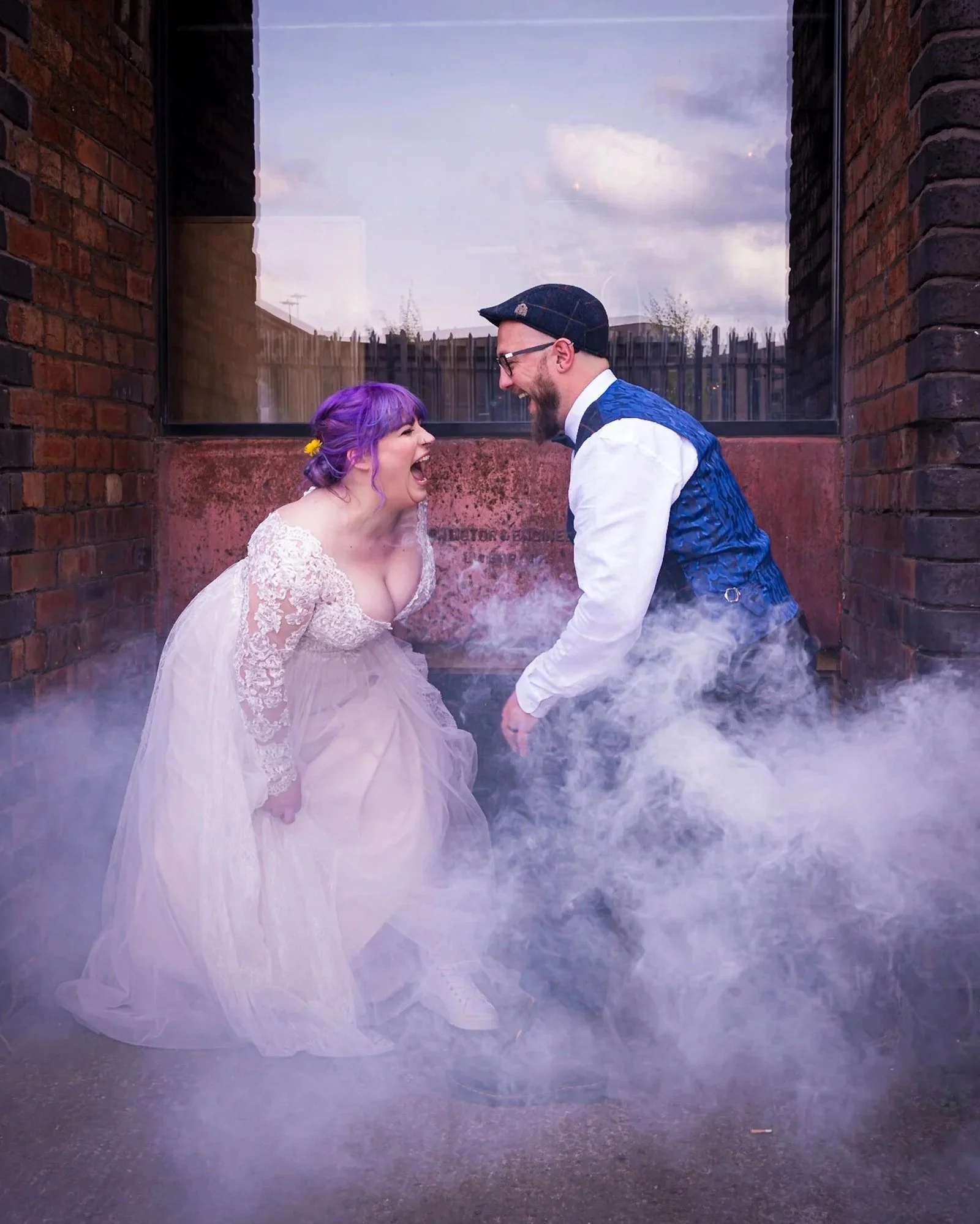 A couple dressed in wedding attire standing close to each other, smiling and laughing in front of a brick wall with a foggy floor and smoke or mist surrounding their feet, under a cloudy sky.